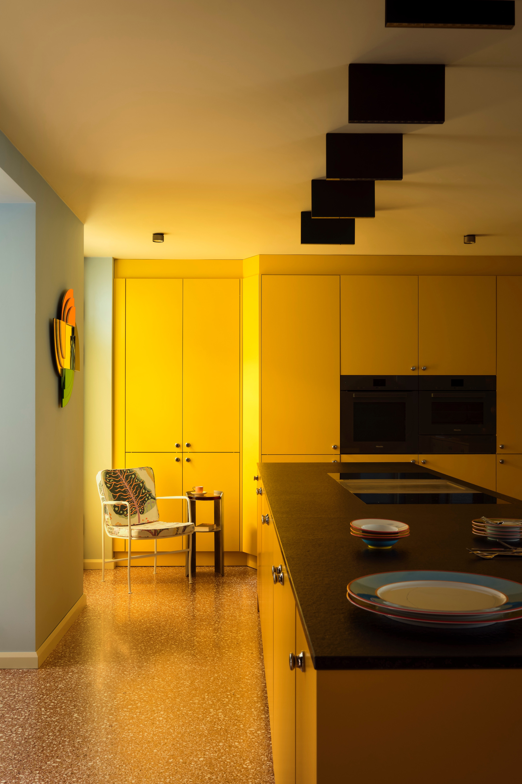 Kitchen with yellow modern units, black worktop, red terrazzo floor and white leaf-patterned armchair