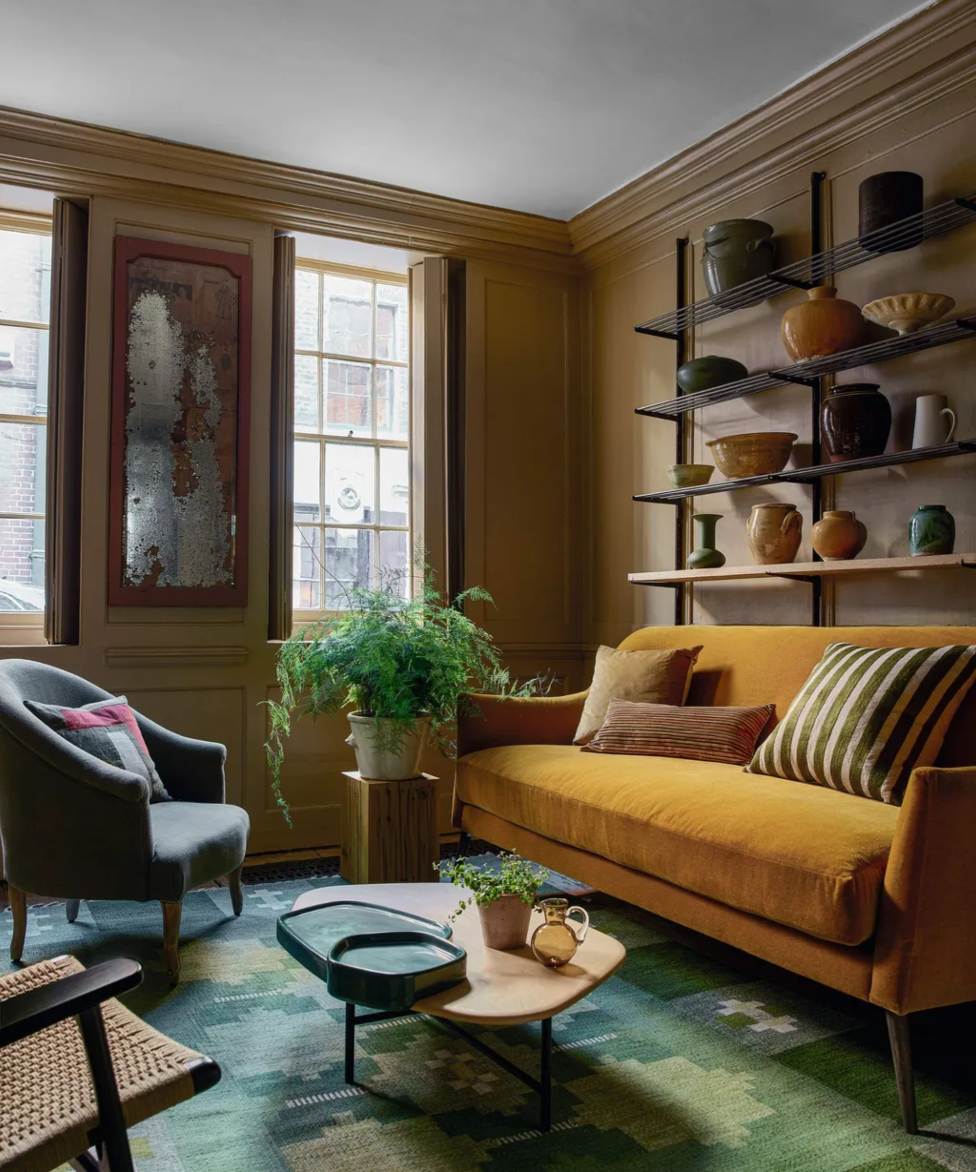 Brown living room with yellow sofa, green rug, and a shelf with pottery hanging from the wall