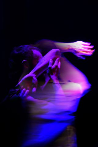The long-exposure photograph of a male ballet dancer caught mid-movement. The background is black and purple and pink light is cast on the dancer.