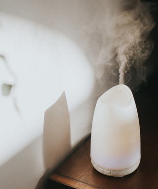 A white essential oil diffuser with must coming out of it on a dark wooden table next to a white mattress