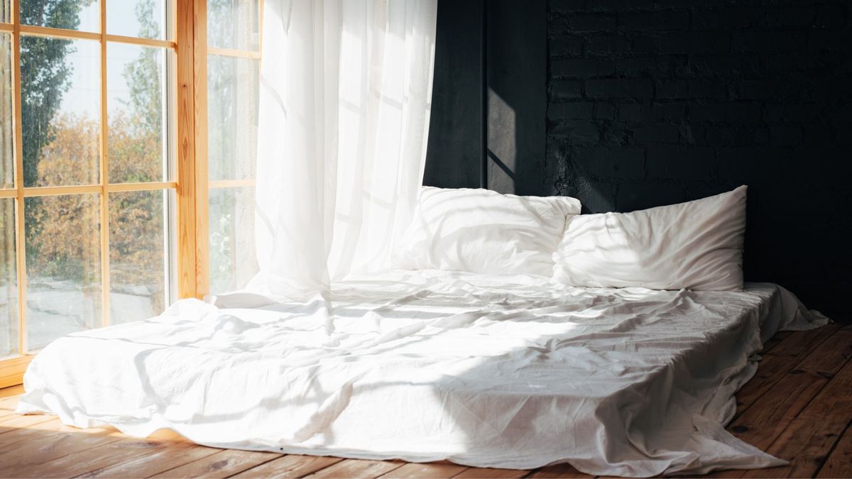 Mattress with white sheets is placed directly on the floor against an open window 