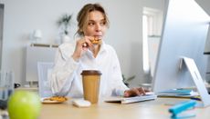 Woman sits at a desk looking at a computer screen. She is eating a chocolate chip cookie from a plate of cookies next to her. A takeaway coffee cup is next to the plate of cookies. A green apple is in the foreground. 