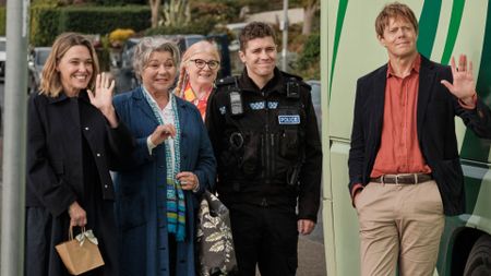 The cast of Beyond Paradise stand and wave by a bus