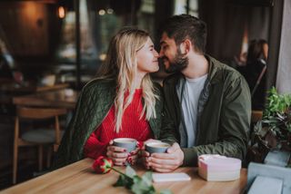 A loved up couple touching noses and smiling while enjoying warm drinks at a cafe.