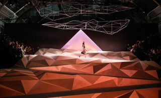 A model walking on a fashion runway designed in the form of mountains in rustic red with audience watching on both sides