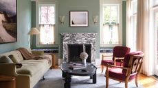 mint green living room with marble fireplace, with artwork and two sconces above it, two red armchairs, upholstered cushion benchseats beneath the two windows either side of the fireplace, a beige sofa with a large artwork above it, two timber side tables and a floor lamp, and a black round coffee table