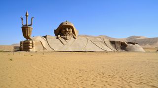 A very tall and wide brownish-colored statue of Genghis Khan in the desert. It shows his upper torso. He is holding a trident coming out of a basket, robes and a hat.