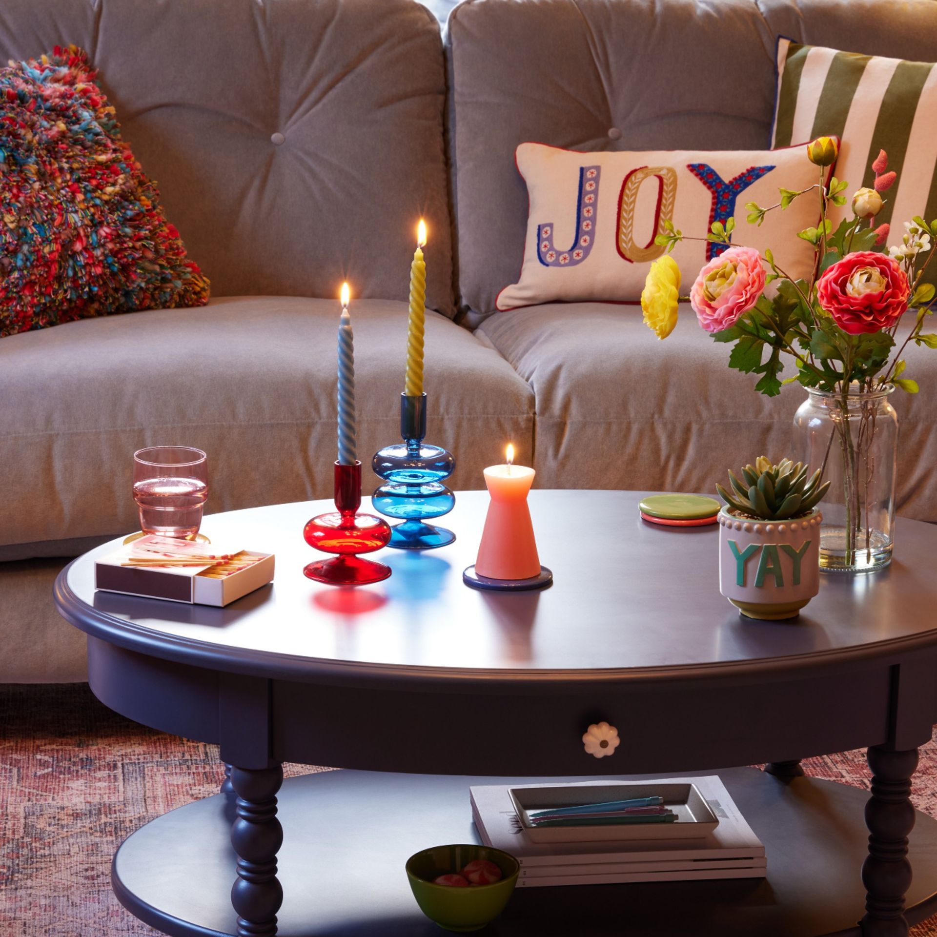 Dunelm Pride and Joy range displayed in a living room, with Joy cushion on a grey sofa and candle holder and plant holder on the table