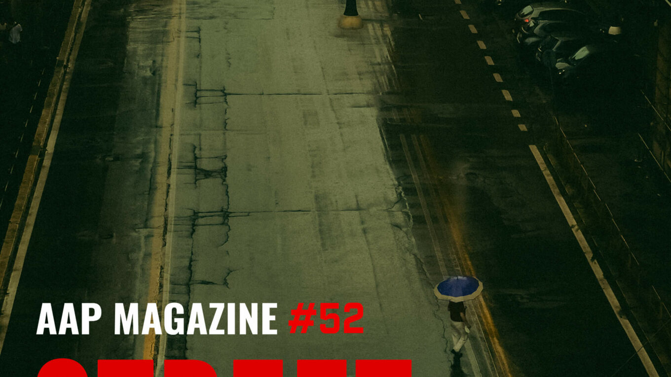 A lone figure with a blue umbrella walks along a wet street, with parked cars lining the road, under a subdued, moody atmosphere