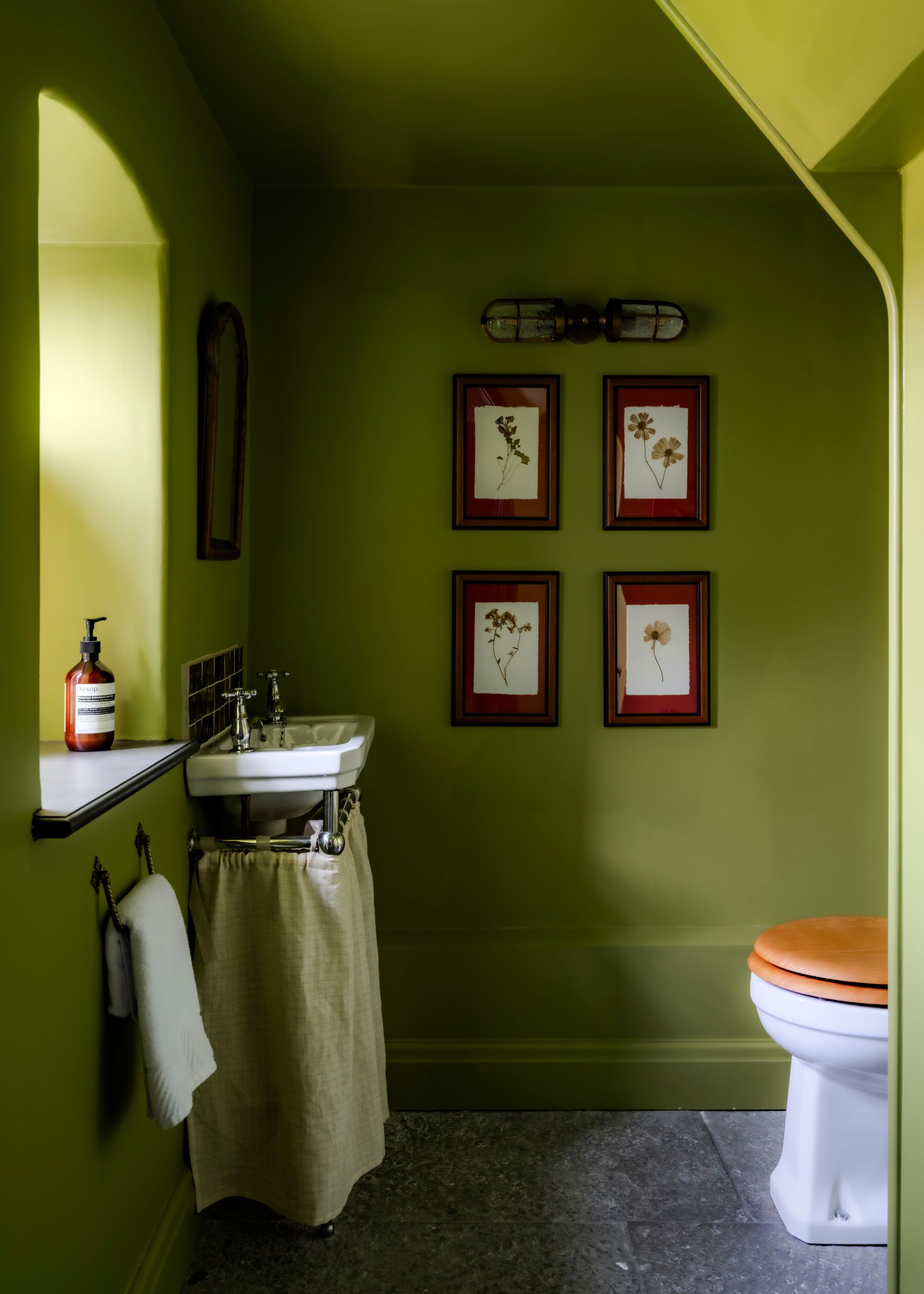 A small bathroom with mossy green color-drenched walls, stone flooring, a small sink with a cafe curtain beneath it and four artworks on the wall.