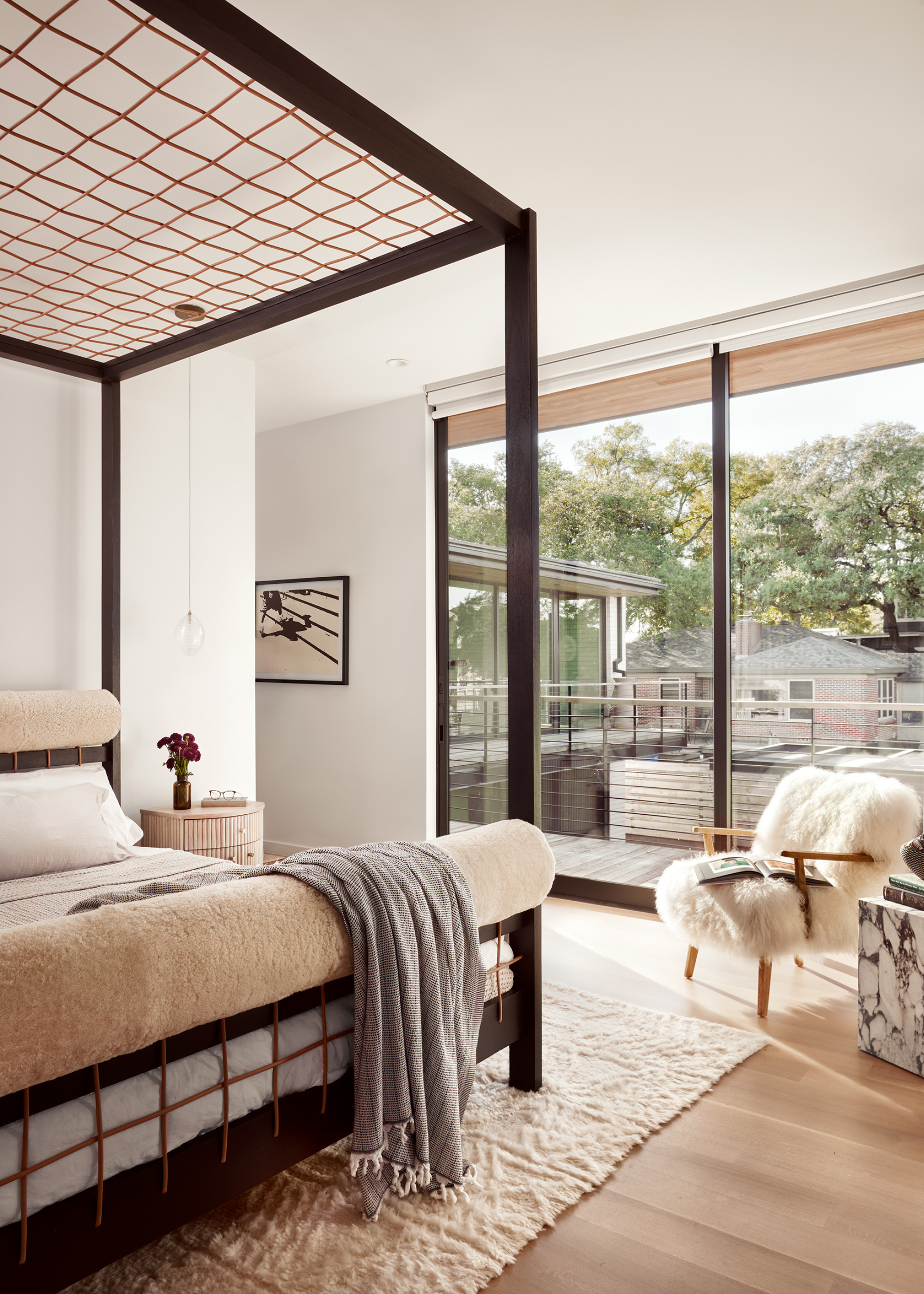 A white bedroom with large windows, a white furry chair, a stone plinth, a side table with a vase of flowers, a hanging bulb, a wood bed frame with a rope design, and a throw blanket