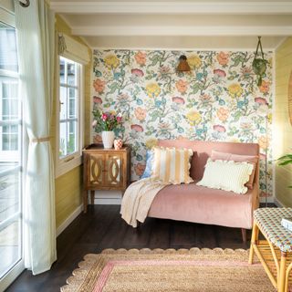 A conservatory living room with a pink sofa bed, a feature wall covered in a floral wallpaper and striped eyelet curtains on the windows