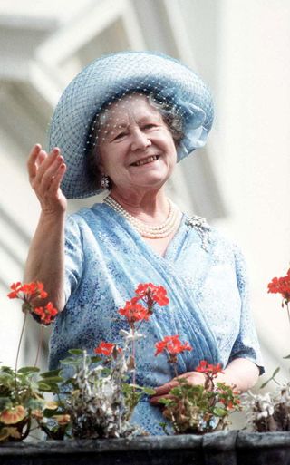 The Queen Mother wearing a blue dress and pearls with a blue hat standing in front of flowers and waving