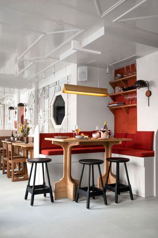 A modernly furnished restaurants features block colored interiors in brown, red, white, and black, including wooden tables, tall stools, art-deco lighting, and more.