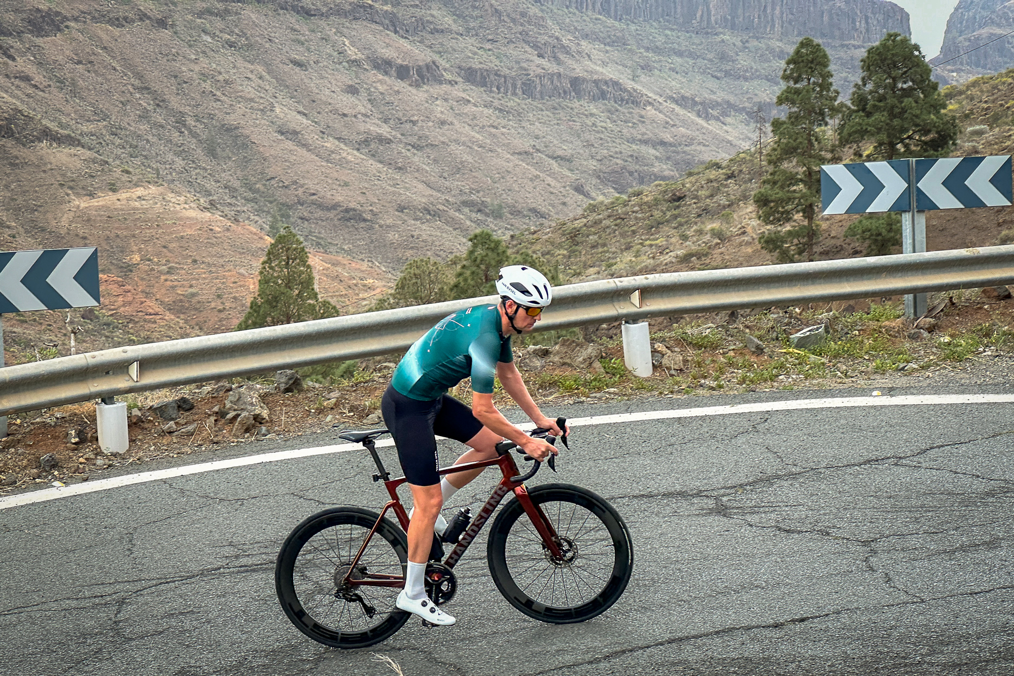 Man riding a road bike up a hill wearing a white helmet, green jersey, black shorts and white shoes.
