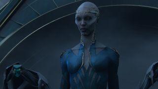 Close up of a gray humanoid alien. They have sharp cheekbones and no hair. They are wearing a simple yet elegant tiara and a blue and silver long-sleeved garment. They are flanked by a robot guard on each side.