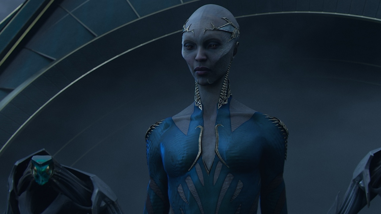 Close by is a gray humanoid alien. He has sharp cheekbones and no hair. She wears a simple but beautiful tiara and a blue and silver sleeve. They are flanked by robot guards on each side.