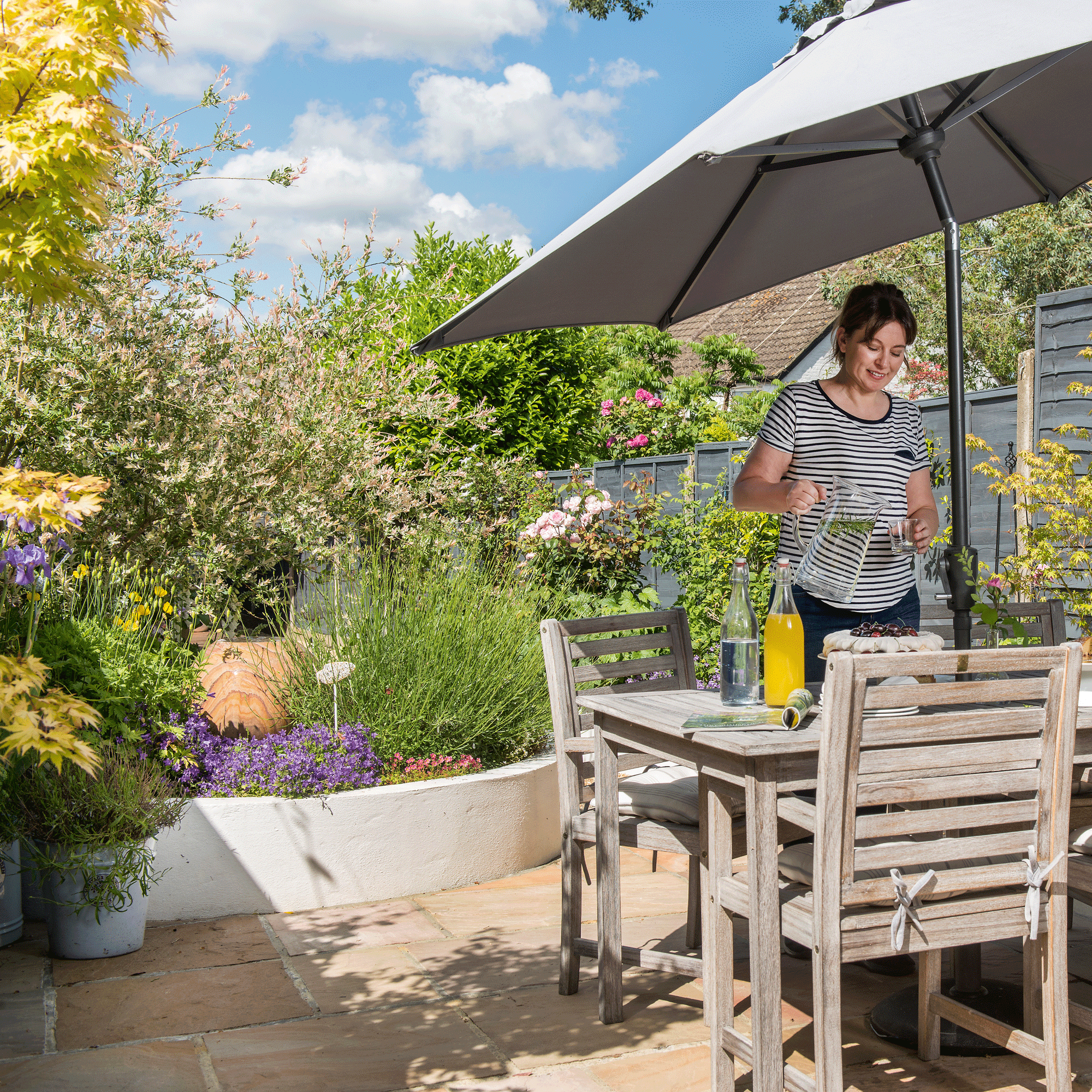 a women in a garden beside a table chairs and parasol surrounded by raised beds and lots of planting and flowers