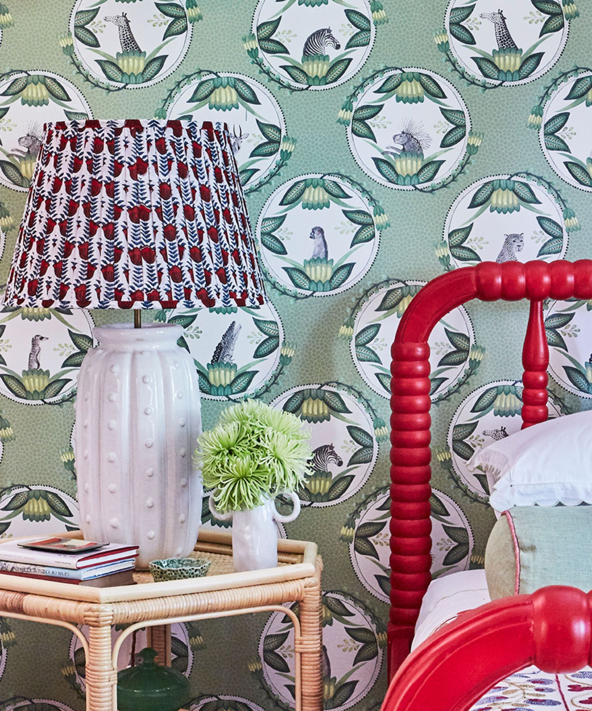 A bright maximalist bedroom with green patterned wallpaper, a red bed frame, and white lamp with clashing pattern shade.