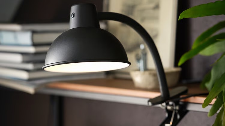 Black IKEA SKURUP clamp spotlight attached to a shelf edge, angled downward in a moody interior