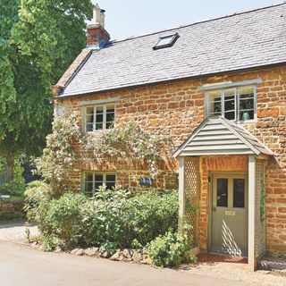 exterior of stone cottage in Cotswolds with wooden canopy porch, slate roof, green paintwork and climbing plants