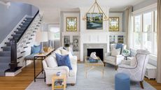 Blue and white living room with a sofa and two armchairs, hanging chandelier, stained glass windows, and miscellaneous lamps and side tables for a maximalist look. A set of stairs is pictured to the right; a bright window is pictured to the left.