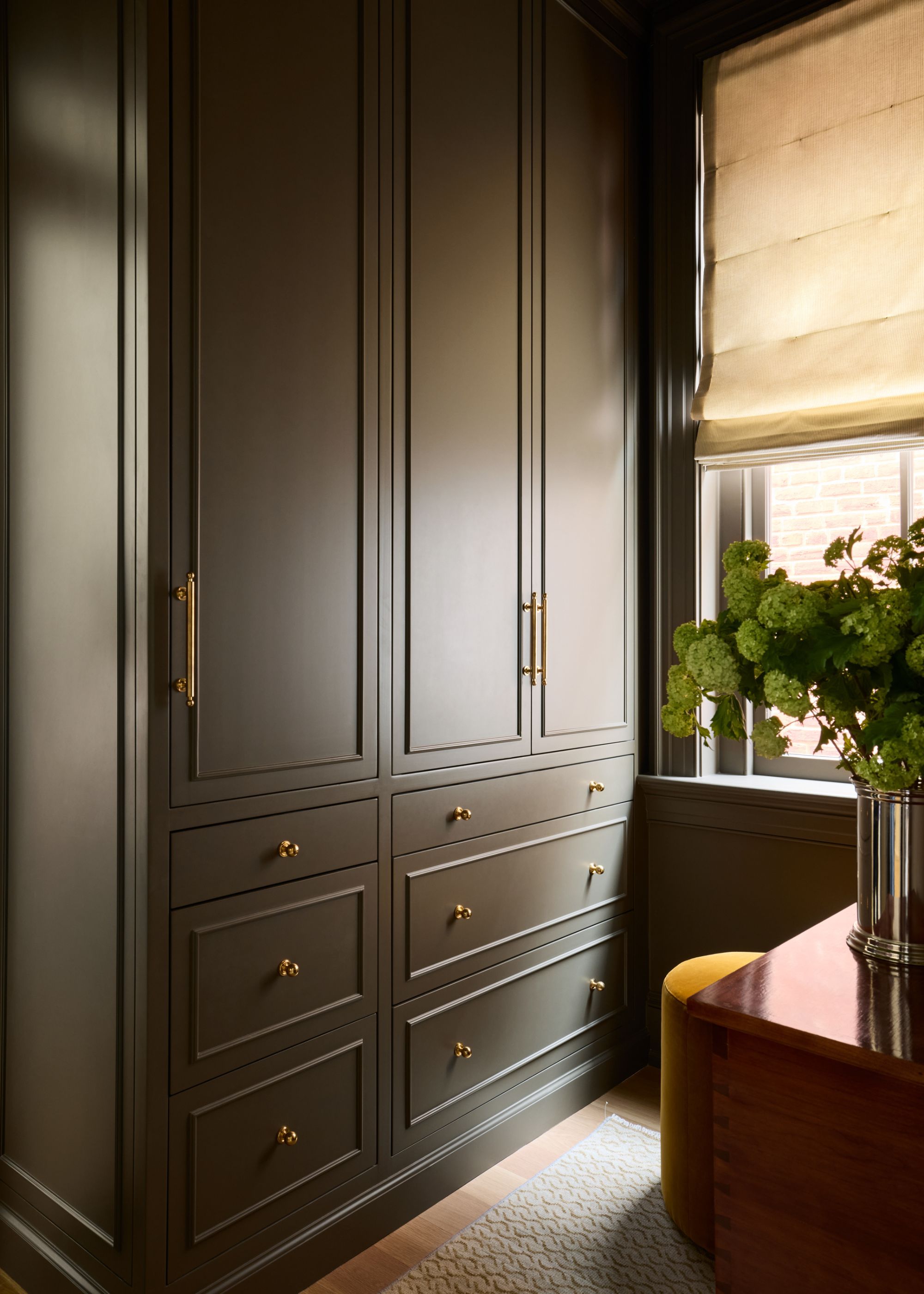 A modern dressing room with full-height cabinetry painted a bronze neutral with brass hardware and a window with a beige Roman blind.