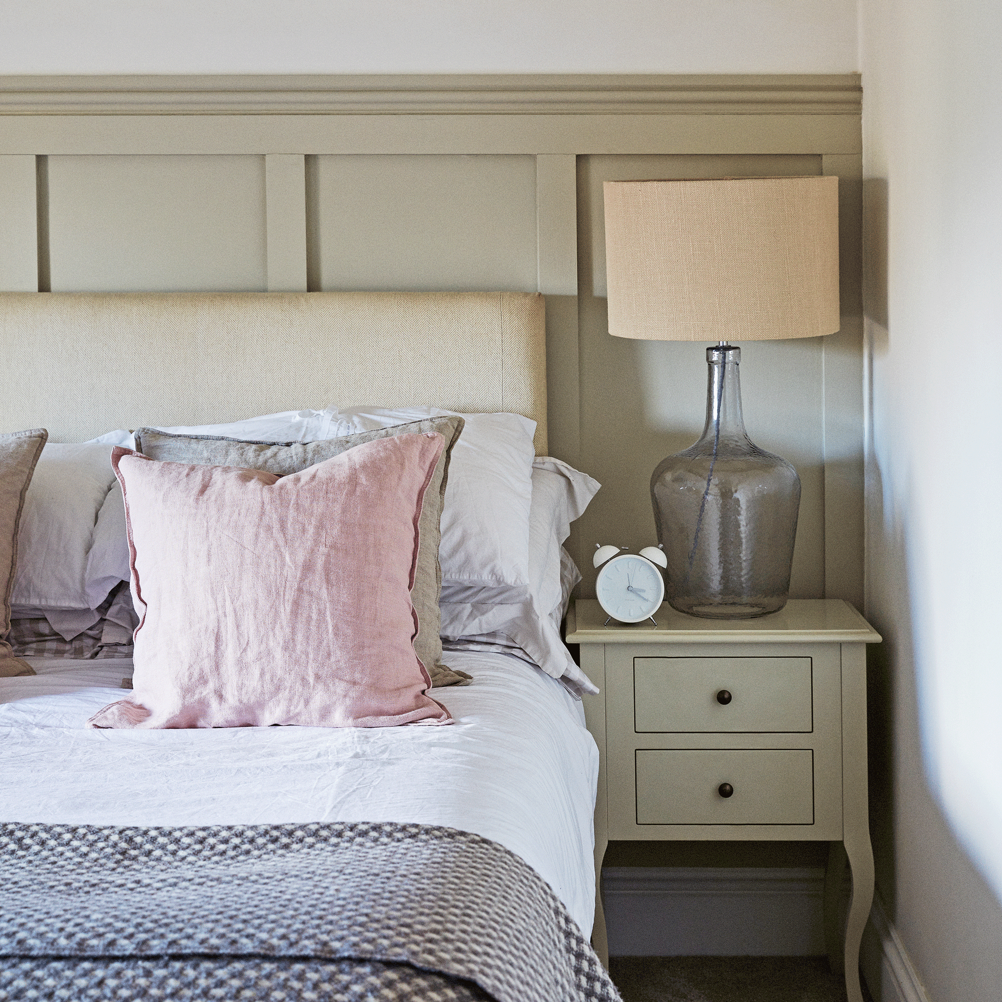 a bedroom with wood panelling beside a bed with layers of bedding and cushions and a large glass-based lamp on the bedside table