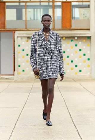 A Chanel model walking in the Resort 2025 show wearing an oversized tweed plaid blazer and patent leather moccasins.