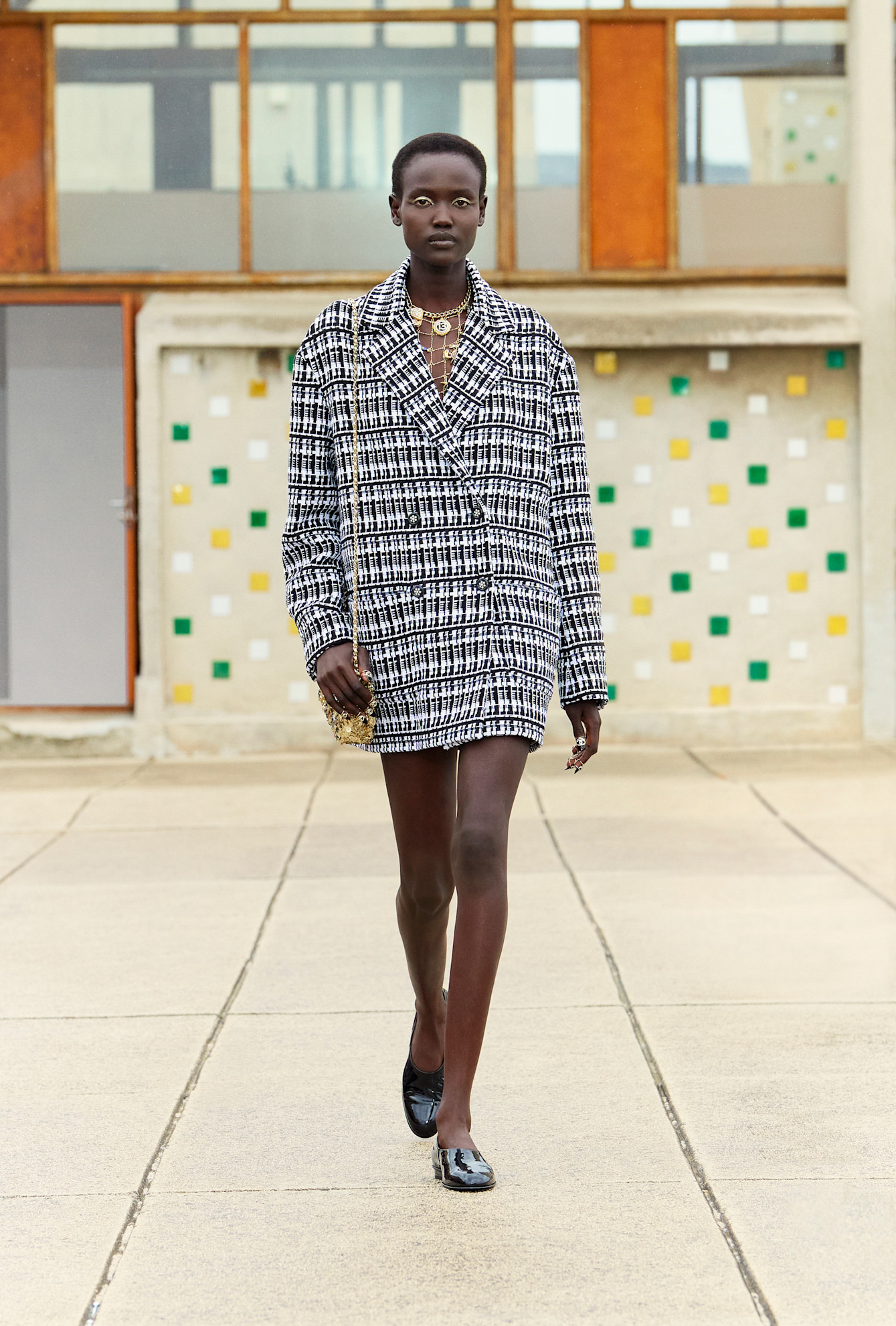 A Chanel model walking in the Resort 2025 show wearing an oversize tweed plaid blazer and patent leather moccasins.