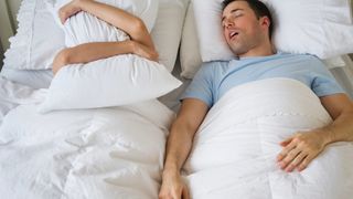 A man with dark hair and wearing a blue shirt snores loudly while sleeping next to his partner, who covers her head with a white pillow to drown out the noise