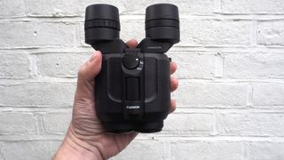 Fujifilm Fujinon Techno-Stabi TS16x28WP binocular on held in a hand in front of a white painted brick wall