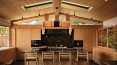 A modern wood kitchen with a pitched roof, a kitchen island with a black countertop and four stools with a large cylindrical pendant light above the island with fringing on either end