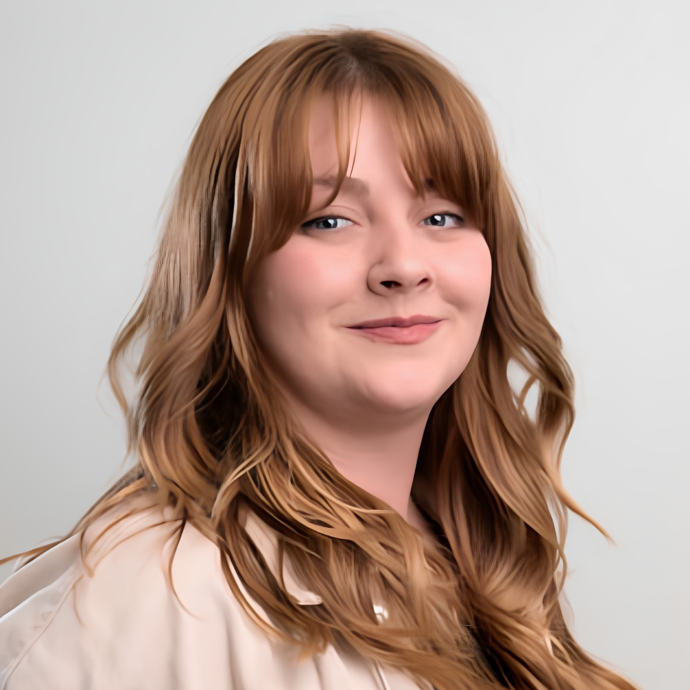 Smiling woman with wavy auburn hair and fringe, wearing a cream jacket, in a studio headshot