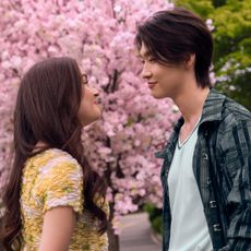 Anna Cathcart as Kitty Song Covey, Sang Heon Lee as Min Ho Moon smiling at each other outside by a cherry blossom tree in xo kitty season 3