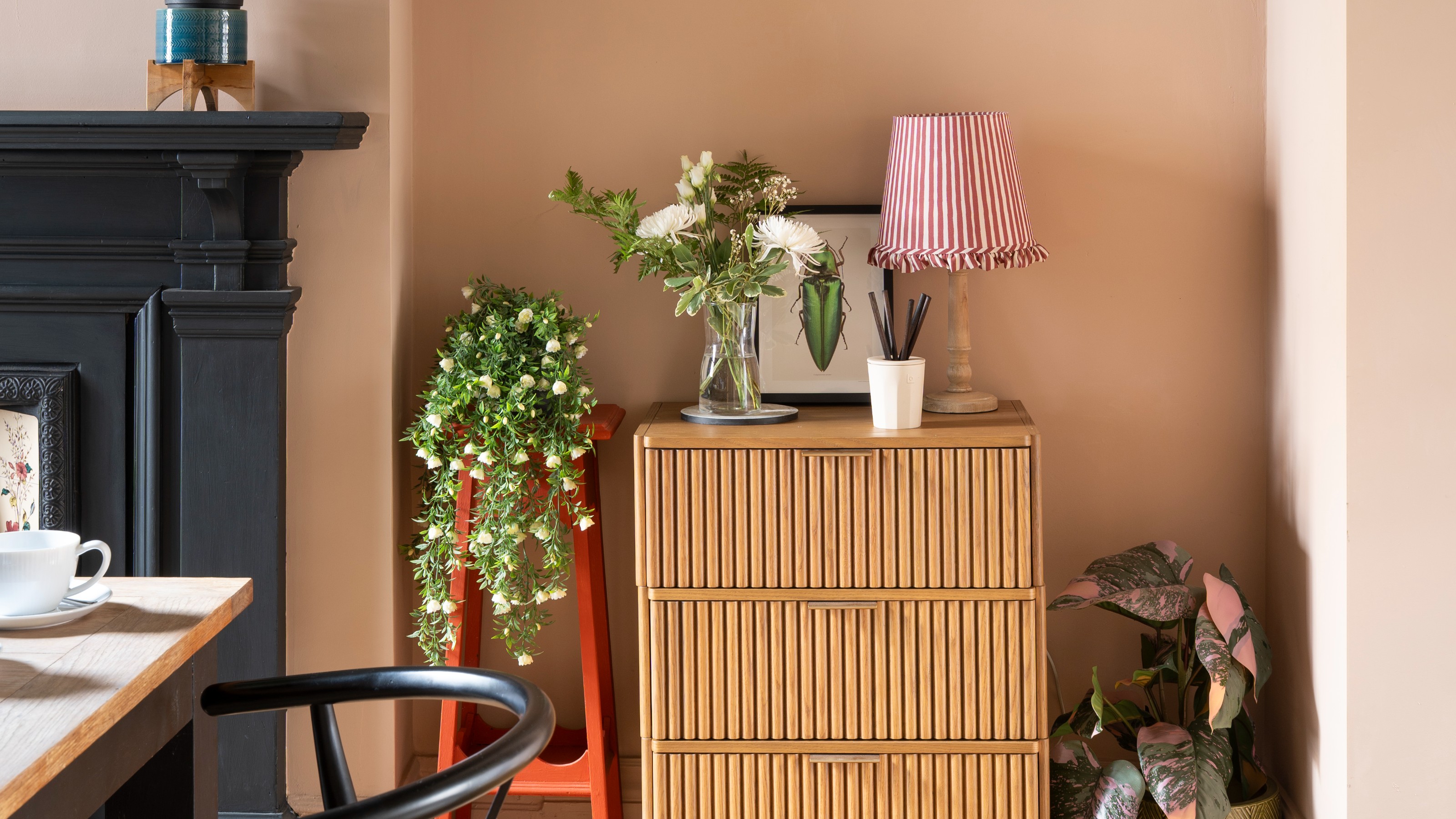 A pink dining room with a black fireplace and a chest of drawers displaying a vase of flowers and a table lamp with a striped shade