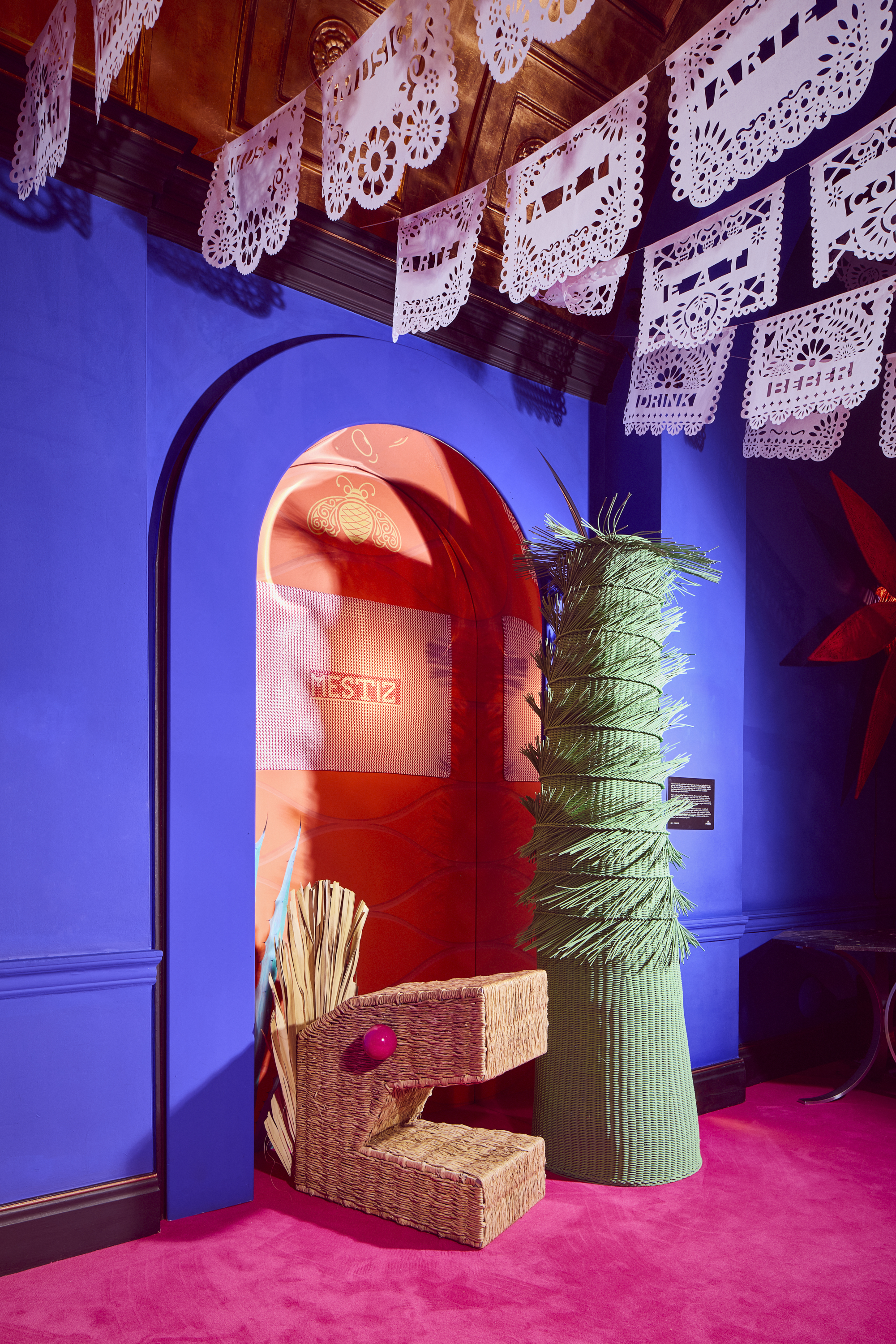 Installation view of a vibrantly colored art show with wicker sculptures resembling cactus, dragons, and sheets of paper bearing different hand-cut messages and hanging from each side of the room.