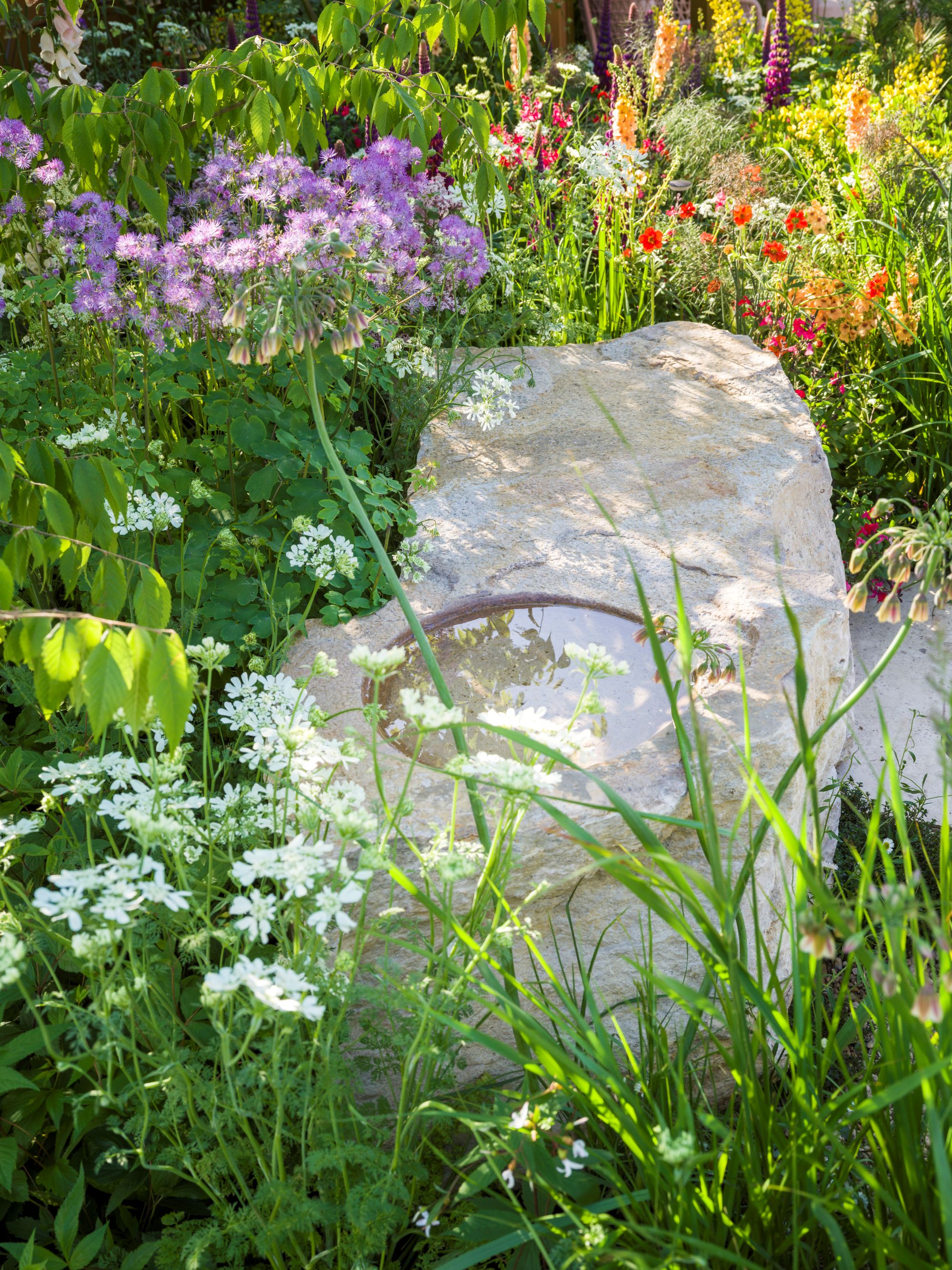 A large pale rock in a garden with a pool of water in the center and surrounded by foliage and colorful flowers