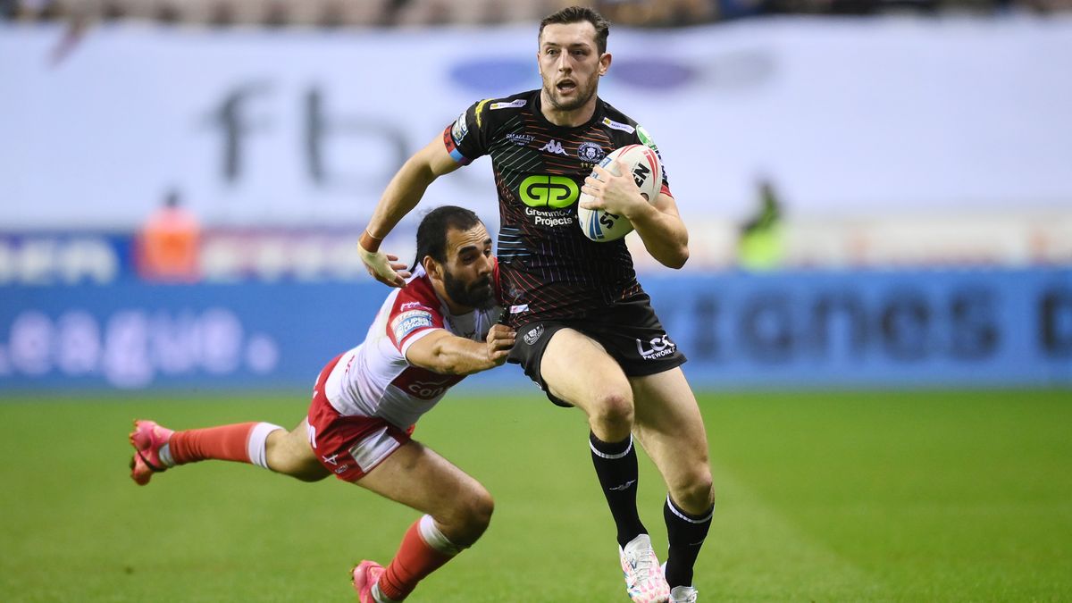 Jake Wardle, in all black kit, skips a challenge from Tyrone May, in red and white, in Wigan vs Hull KR.