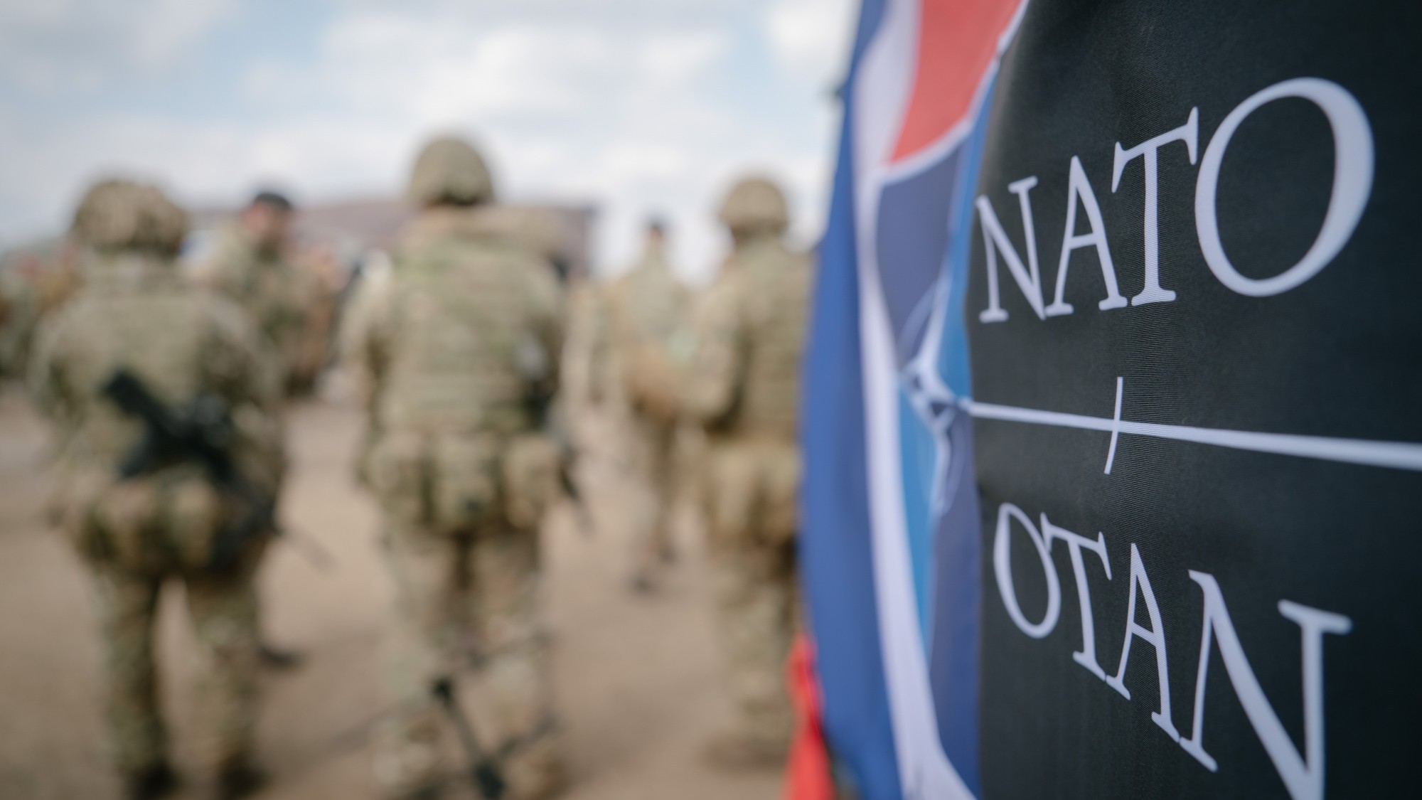 Close up of a Nato logo, with blurred soldiers in the background