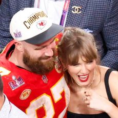 Travis Kelce wears his Kansas City Chiefs uniform and a baseball cap as he hugs girlfriend Taylor Swift on the field after the Super Bowl