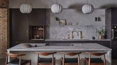 Three silk pendant lantern lights hanging over a white marble kitchen island there is dark gray cabinetry and four wooden bar stools