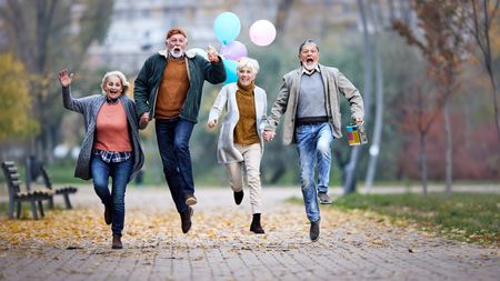 Group of playful mature friends having fun while jumping with balloons in autumn day at the park. Copy space.