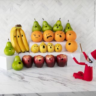 an elf conducting fruit on the kitchen counter, with the fruit having had faces drawn onto them with pen