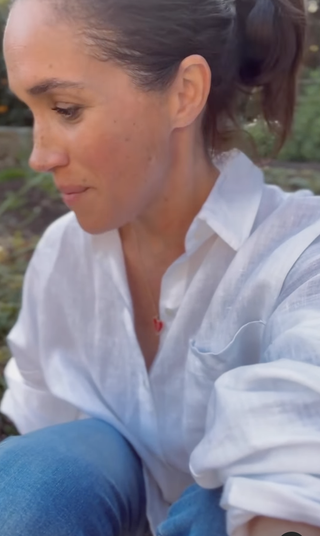 Meghan Markle wearing a white blouse and jeans turned to the side kneeling in a garden