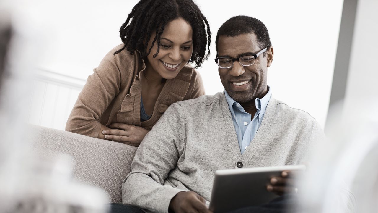 A woman looks over a man&#039;s shoulder as they look at his tablet together.