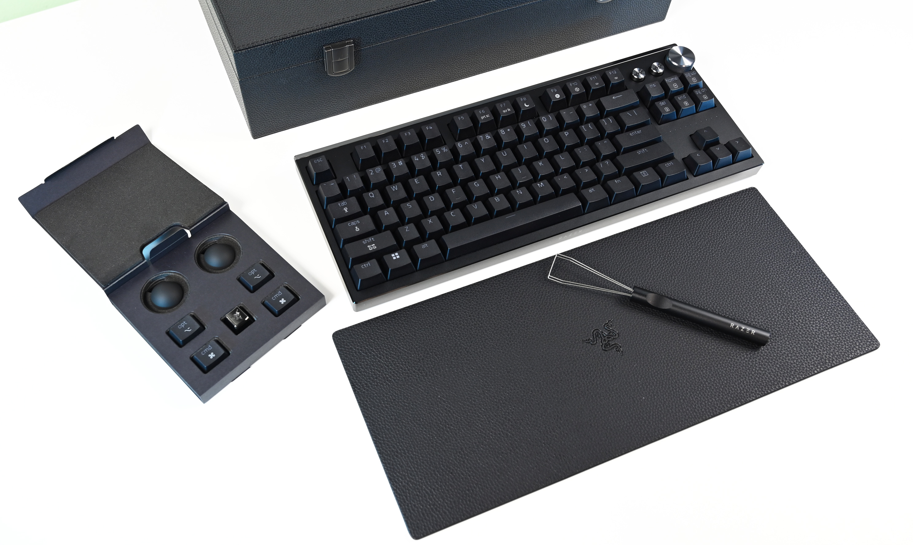 Overhead view of the keyboard, a textured leather-style desk mat, a wire keycap puller, an extra keycap tray, and the premium storage box on a white surface.