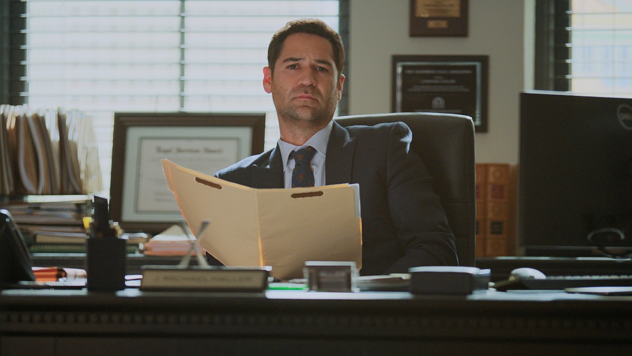 Manuel Garcia-Rulfo sitting behind a desk and holding up a file in The Lincoln Lawyer. 