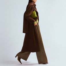 Model wearing a long brown coat and brown wide leg trousers with a lime green handbag and top walking in a studio for Harvey Nichols SS25