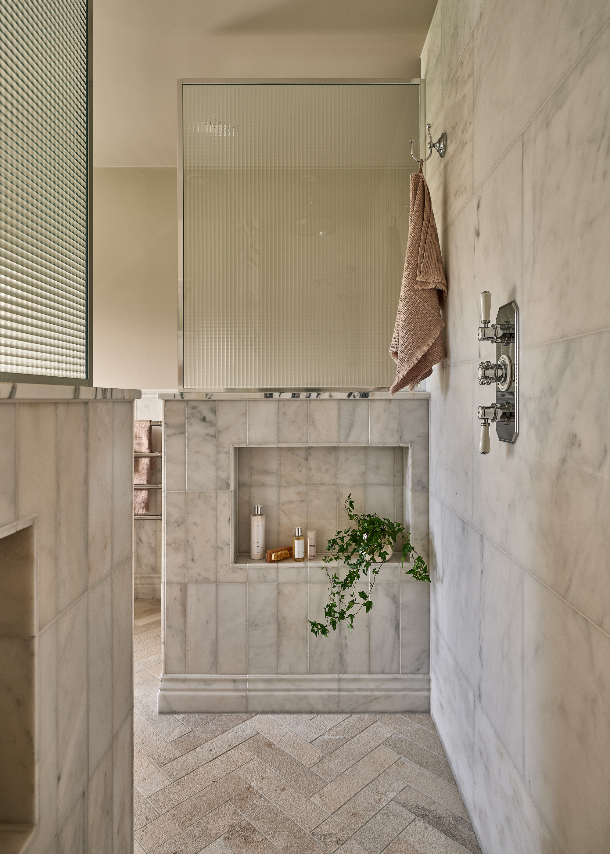 a large walk in shower with matt, honed marble tiles, a cubby hole with plants in it, and half glass walls